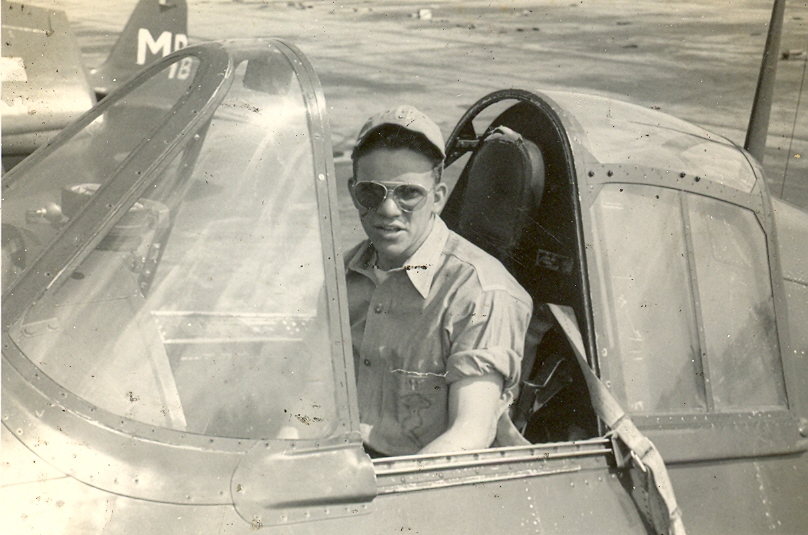 Bob Neely, wearing sunglasses and a cap, sits in the cockpit of a Grumman F6F Hellcat on a tarmac. The plane’s canopy is open, and another aircraft tail is visible in the background. The photo has a sepia tone.