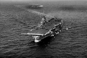 Aerial view of a large aircraft carrier navigating through the ocean, leaving a visible wake behind it on a calm sea.