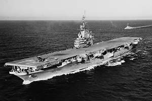 Black and white aerial photo of the USS Oriskany, a large aircraft carrier, sailing on the ocean with several airplanes on its deck. Another vessel follows in the distance. The sea is calm, and the sky is clear.