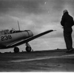 A vintage black and white photo shows a person wearing a helmet standing on an aircraft carrier deck as a military propeller plane marked with the number 238 prepares for takeoff. The sky is overcast with clouds.